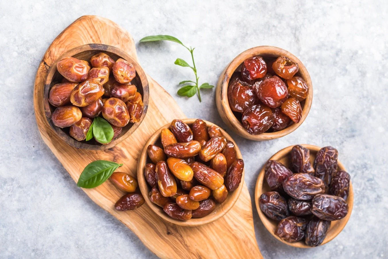 Empat mangkuk kayu berisi kurma yang berlainan warna dan bentuk, daripada keemasan terang hingga coklat gelap, disusun di atas papan kayu.